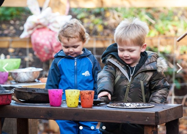 Two young children playing with cups in an outdoor mud kitchen