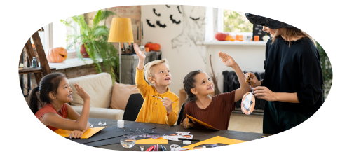Three children and an adult gathered around a table with crafting materials. The adult is holding a Halloween decoration while the children hold up their hands. A pumpkin and several bat decorations are visible in the background.