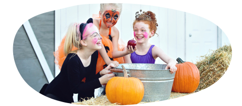Three children dressed in Halloween costumes standing around a large bucket. All three children have face paint on and are laughing. Two pumpkins surrounf