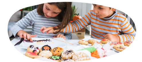 Two children sitting at a table decorated with various colourful cookies. One child is reaching for a cooking while the other focuses on decorating some paper with dark icing.