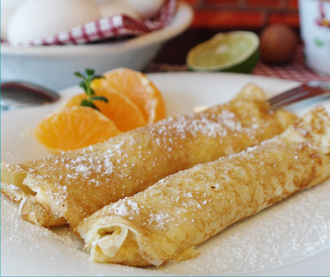 Thin pancakes rolled up on a white plate, dusted with powdered sugar, and sitting beside 3 segments of satsuma